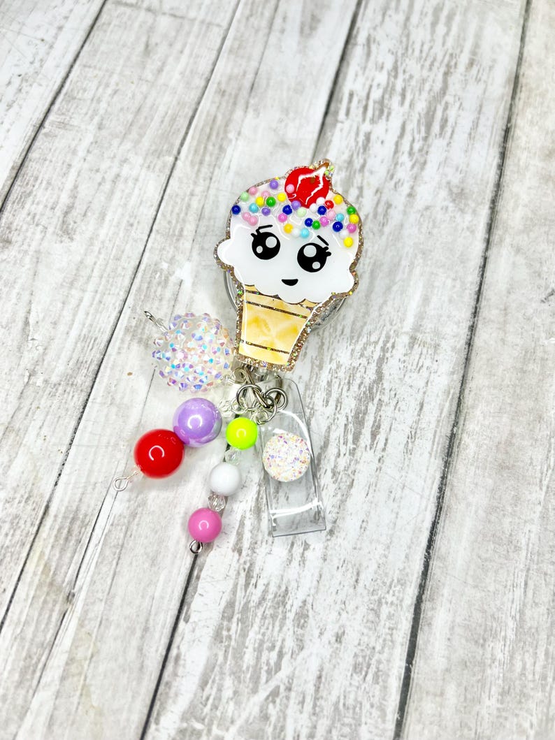 Op de afbeelding: Een decoratieve badgehaspel in de vorm van een ijsje met een lachend gezicht, kleurrijke hagelslag en een rode kers erop. De badgehaspel heeft een doorzichtige plastic ID-houder en een kralenbedel.
