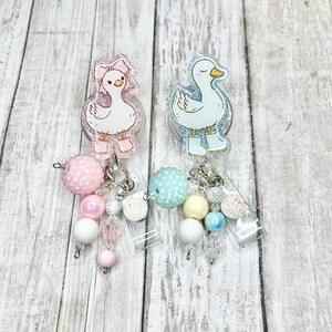 Puede incluir: Dos portatarjetas de acr&iacute;lico con dise&ntilde;os de patos de dibujos animados. Un pato lleva un lazo y botas rosas, el otro botas azules. Cada carrete tiene un dije de cuentas con cuentas rosas, blancas y pastel. El fondo es de vetas de madera clara.