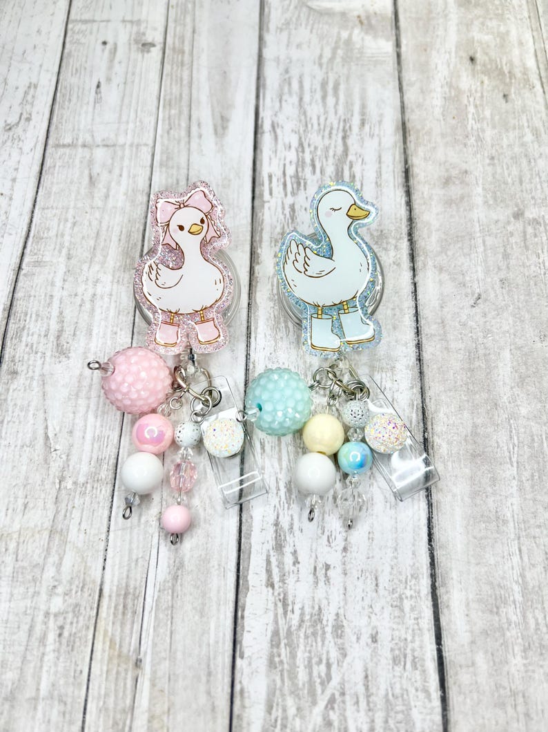 Puede incluir: Dos portatarjetas de acr&iacute;lico con dise&ntilde;os de patos de dibujos animados. Un pato lleva un lazo y botas rosas, el otro botas azules. Cada carrete tiene un dije de cuentas con cuentas rosas, blancas y pastel. El fondo es de vetas de madera clara.