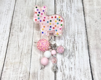 May include: A pink, glittery badge reel in the shape of a horse, adorned with colorful dots. A dangling charm features pink and white beads, a glitter ball, and clear accents. The reel is set against a light wood grain background.