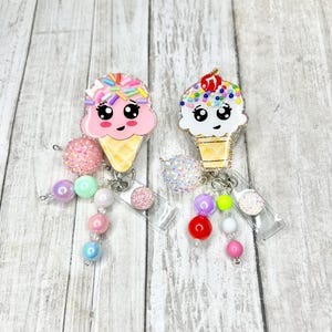 Op de afbeelding: Twee badge-haspels in de vorm van een ijshoorntje. E&eacute;n is roze met regenboogkleurige hagelslag, de andere is wit met kleurrijke hagelslag en een kers. Elk heeft een kralenbedel met diverse pastelkleurige en glinsterende kralen.