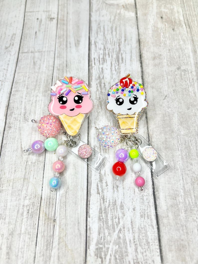 Op de afbeelding: Twee badge-haspels in de vorm van een ijshoorntje. E&eacute;n is roze met regenboogkleurige hagelslag, de andere is wit met kleurrijke hagelslag en een kers. Elk heeft een kralenbedel met diverse pastelkleurige en glinsterende kralen.