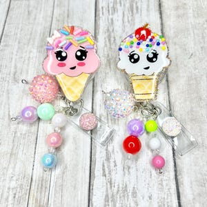 Op de afbeelding: Twee badgehaspels in de vorm van een ijshoorntje. De ene is roze met regenboogkleurige sprinkles, de andere is wit met kleurrijke sprinkles en een kers. Elk heeft een kralenbedel met pastelkleurige en glinsterende kralen.