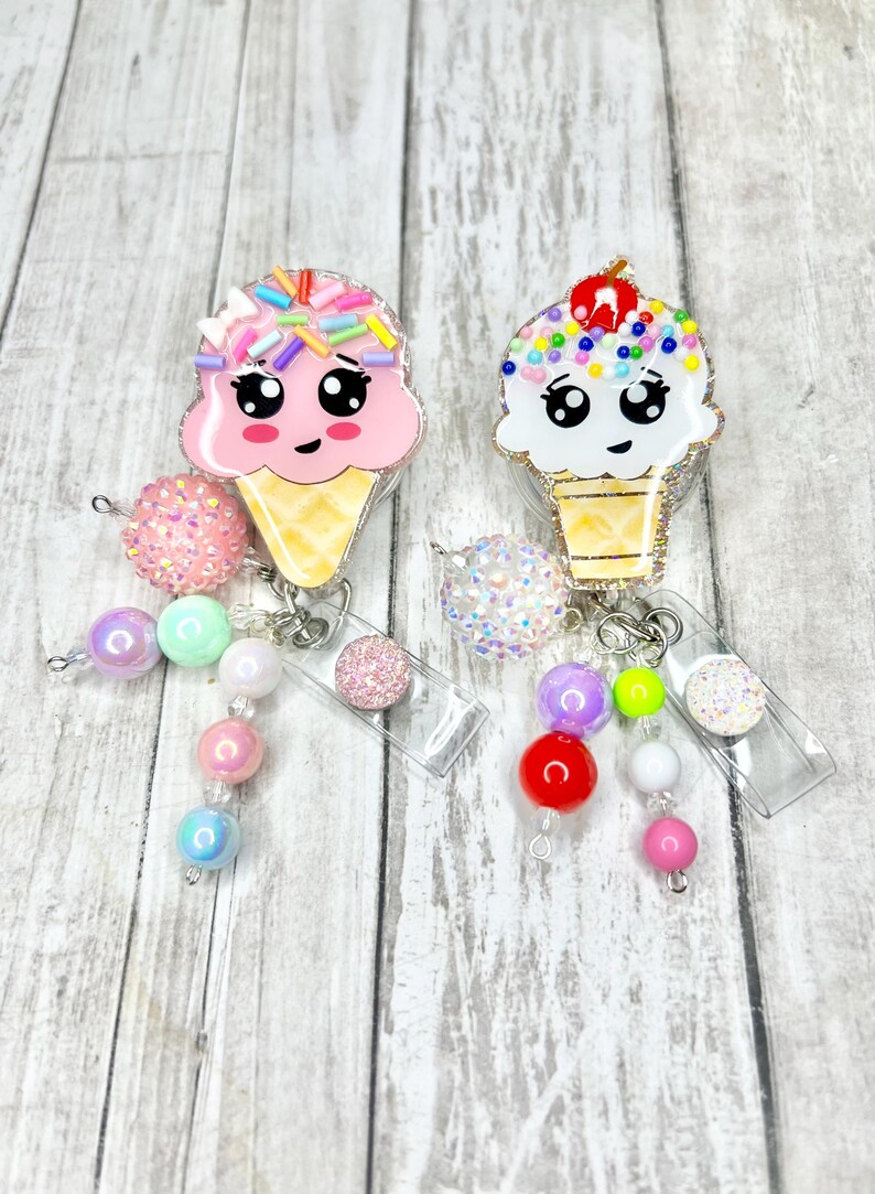 Op de afbeelding: Twee badgehaspels in de vorm van een ijshoorntje. De ene is roze met regenboogkleurige sprinkles, de andere is wit met kleurrijke sprinkles en een kers. Elk heeft een kralenbedel met pastelkleurige en glinsterende kralen.