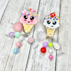 Op de afbeelding: Twee badgehaspels in de vorm van ijsjes met lachende gezichten. De ene is roze met kleurrijke sprinkles, de andere wit met een kers erop. Elk heeft een kralenbedel met roze, paarse en iriserende kralen.