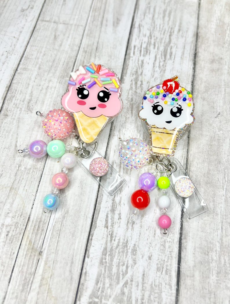 Op de afbeelding: Twee badgehaspels in de vorm van ijsjes met lachende gezichten. De ene is roze met kleurrijke sprinkles, de andere wit met een kers erop. Elk heeft een kralenbedel met roze, paarse en iriserende kralen.