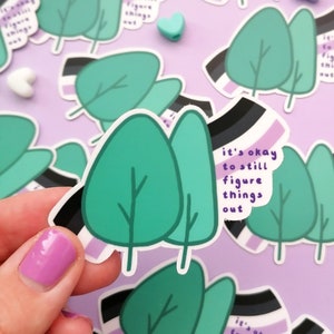 Könnte beinhalten: Ein Set von Aufklebern mit einer stilisierten Illustration von drei Bäumen mit dem Text "it's okay to still figure things out" in lila und schwarzer Schrift auf weißem Hintergrund. Die Aufkleber haben ein lila, schwarzes und weißes Streifenmuster.