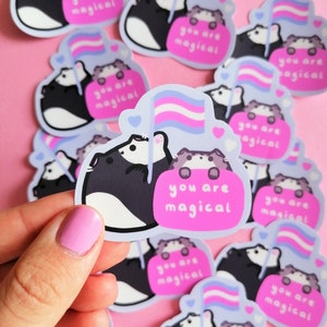 Op de afbeelding: Een set stickers met twee cartoondieren die een transgender pride-vlag vasthouden. De stickers zijn op een roze achtergrond en elke sticker zegt "Je bent magisch".