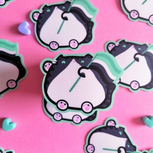 May include: A set of stickers featuring a cartoon cat holding a green, black, and white pride flag. The cat is white with black markings and has pink paws. The stickers are on a pink background.