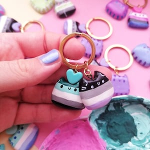 May include: Two hand-painted cat keychains with a heart charm. The cats are painted in the colours of the ace flag, with black, white, grey, purple, and light blue stripes. The keychains are held in a hand with light blue nail polish.