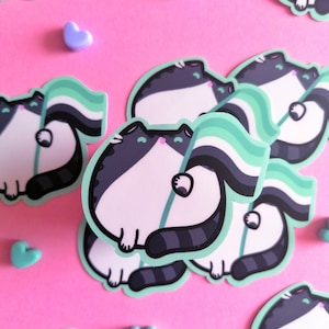 May include: A set of stickers featuring a cartoon cat holding a green, black, and white pride flag. The cat is white with black stripes and has a pink nose. The stickers are on a pink background.