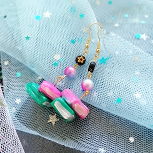 May include: A pair of dangle earrings with a pink, green and white geometric design. The earrings are made of polymer clay and have gold-coloured hooks. The earrings are hanging on a blue tulle fabric with white stars.