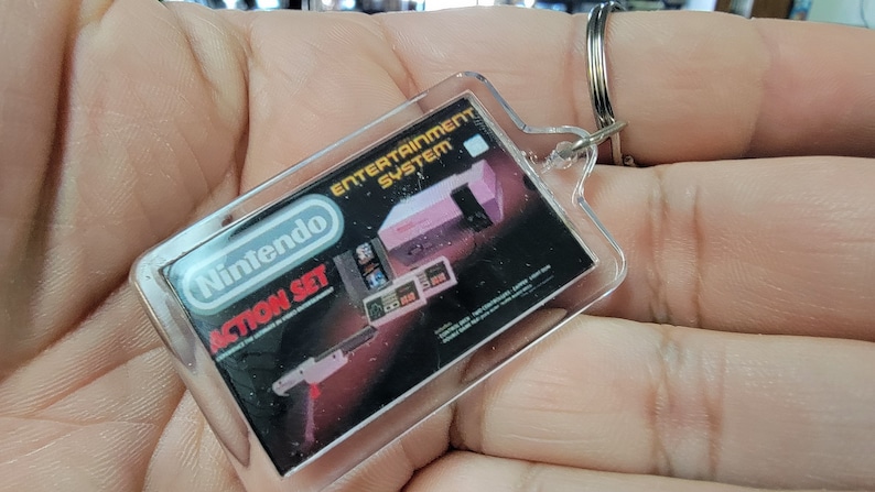 May include: A clear plastic keychain with a black and white image of a Nintendo Entertainment System Action Set. The image includes the text "Nintendo Entertainment System" and "Action Set".