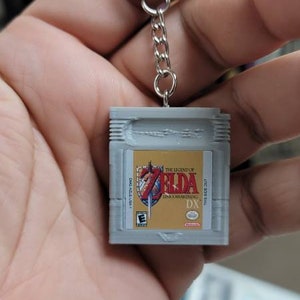 May include: A grey and gold keychain shaped like a Game Boy Color cartridge featuring the game "The Legend of Zelda: Link's Awakening DX".