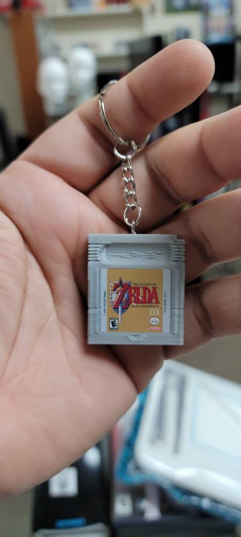 May include: A grey and gold keychain shaped like a Game Boy Color cartridge featuring the game "The Legend of Zelda: Link's Awakening DX".