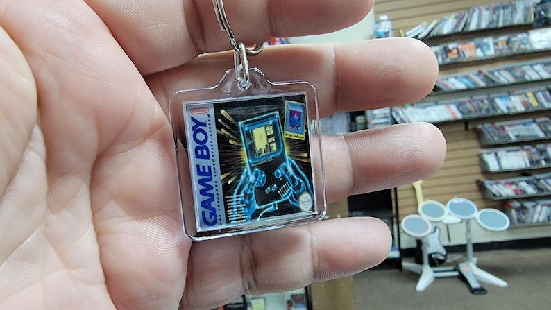 May include: A clear plastic keychain featuring a Game Boy Color with a blue and black design. The keychain has a silver metal ring.