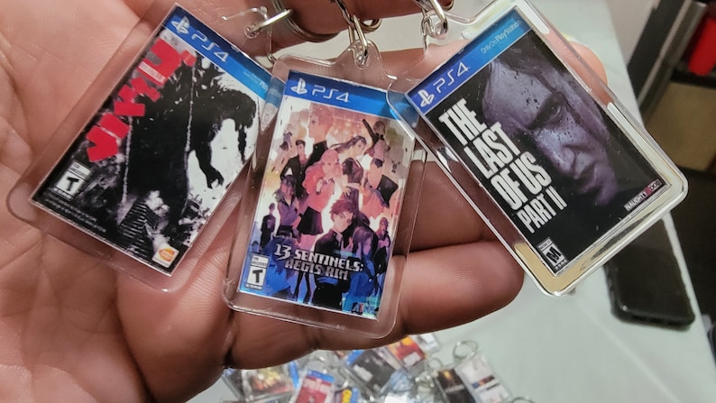 Puede incluir: Tres llaveros de juegos de PlayStation 4 con las portadas de "The Last of Us Part II", "13 Sentinels: Aegis Rim" y "Godzilla".