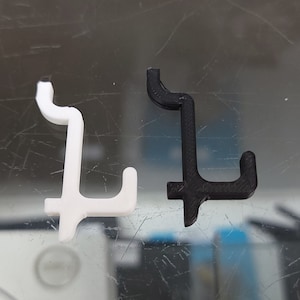 May include: Two plastic hooks, one white and one black, are shown on a reflective surface. The hooks are shaped like the letter L.