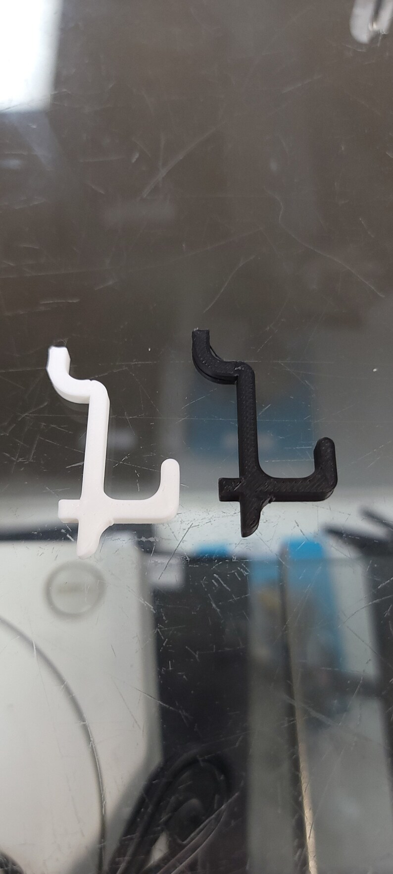 May include: Two plastic hooks, one white and one black, are shown on a reflective surface. The hooks are shaped like the letter L.