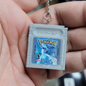 May include: A grey plastic keychain shaped like a Game Boy Color game cartridge. The cartridge has a label with the text "Pok&eacute;mon" and a picture of the Pok&eacute;mon Lugia. The label also says "Silver Version".