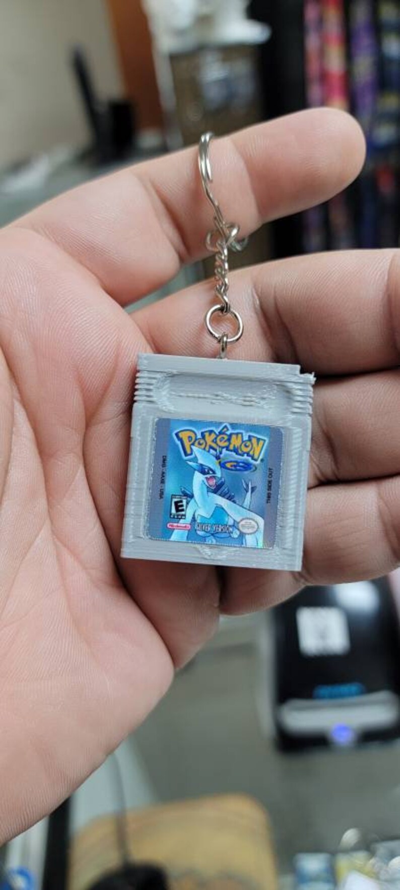 May include: A grey plastic keychain shaped like a Game Boy Color game cartridge. The cartridge has a label with the text "Pok&eacute;mon" and a picture of the Pok&eacute;mon Lugia. The label also says "Silver Version".