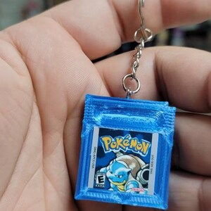May include: A blue Game Boy Color keychain with a Pokemon game cartridge design. The cartridge features a blue and white image of Squirtle, a water-type Pokemon.