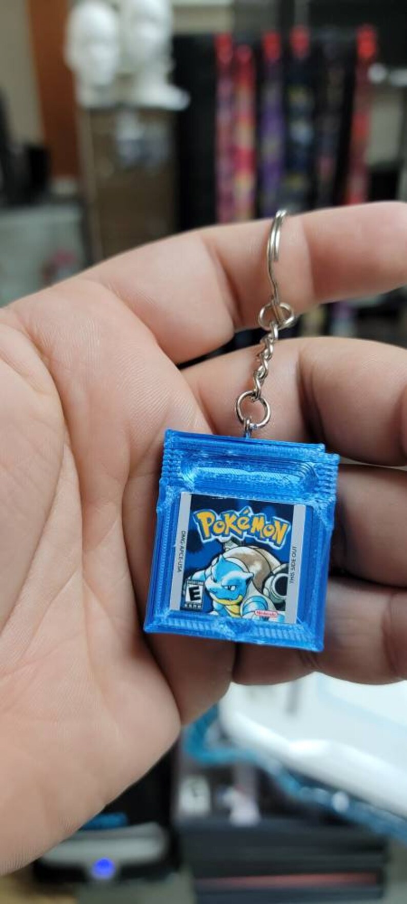 May include: A blue Game Boy Color keychain with a Pokemon game cartridge design. The cartridge features a blue and white image of Squirtle, a water-type Pokemon.