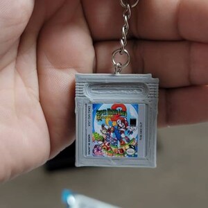 May include: A silver Game Boy Color keychain with a Super Mario Land 2: 6 Golden Coins game cartridge design. The keychain features a silver chain and a silver ring.