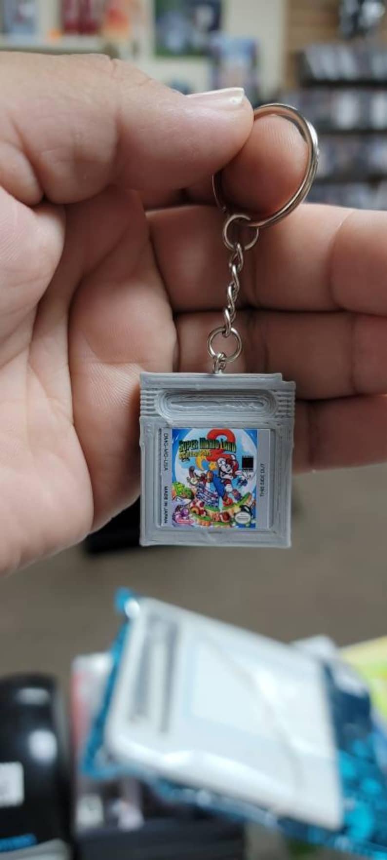 May include: A silver Game Boy Color keychain with a Super Mario Land 2: 6 Golden Coins game cartridge design. The keychain features a silver chain and a silver ring.
