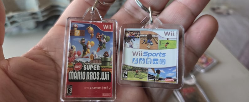 K&ouml;nnte beinhalten: Zwei durchsichtige Schl&uuml;sselanh&auml;nger aus Kunststoff mit Bildern von Nintendo Wii-Videospiel-H&uuml;llen. Der linke Schl&uuml;sselanh&auml;nger zeigt das Cover von "New Super Mario Bros. Wii" und der rechte Schl&uuml;sselanh&auml;nger zeigt das Cover von "Wii Sports".