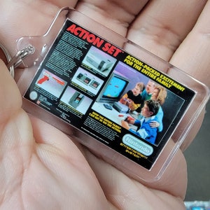 May include: A clear plastic keychain with a black and white image of a Nintendo Entertainment System Action Set. The image shows a family playing the NES with the text "Action-Packed Excitement for the Entire Family".