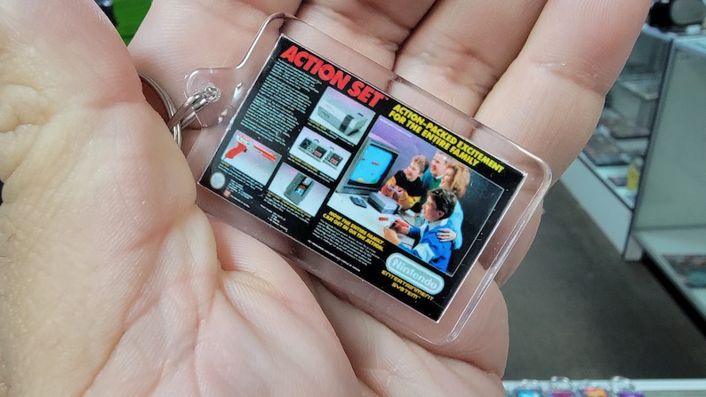 May include: A clear plastic keychain with a black and white image of a Nintendo Entertainment System Action Set. The image shows a family playing the NES with the text "Action-Packed Excitement for the Entire Family".