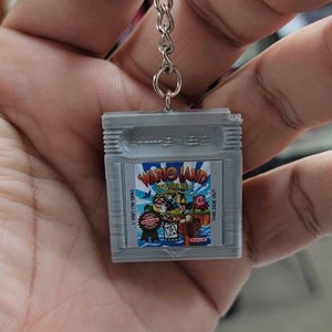 May include: A grey Game Boy Color keychain featuring a Wario Land Super Mario Land game cartridge design. The keychain has a silver metal chain and ring.
