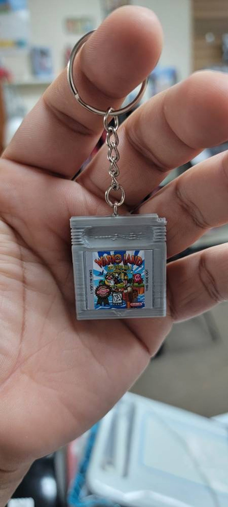 May include: A grey Game Boy Color keychain featuring a Wario Land Super Mario Land game cartridge design. The keychain has a silver metal chain and ring.