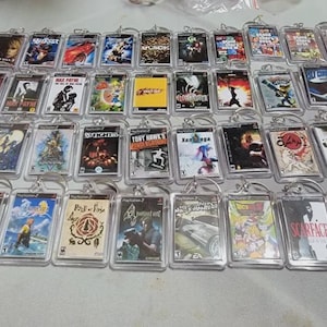 May include: A collection of video game keychains featuring various game covers. The keychains are encased in clear plastic and attached to silver keyrings. The game covers display colorful graphics and titles, showcasing a variety of game genres.