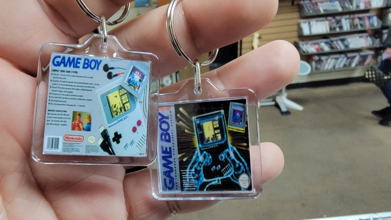 May include: Two clear plastic keychains featuring images of the Game Boy handheld video game console. The first keychain shows a white Game Boy with blue and white box art. The second keychain shows a blue and green Game Boy with black and white box art.
