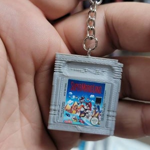 May include: A grey Game Boy Color cartridge keychain with a Super Mario Land game label. The keychain has a silver metal chain attached to a silver metal ring.