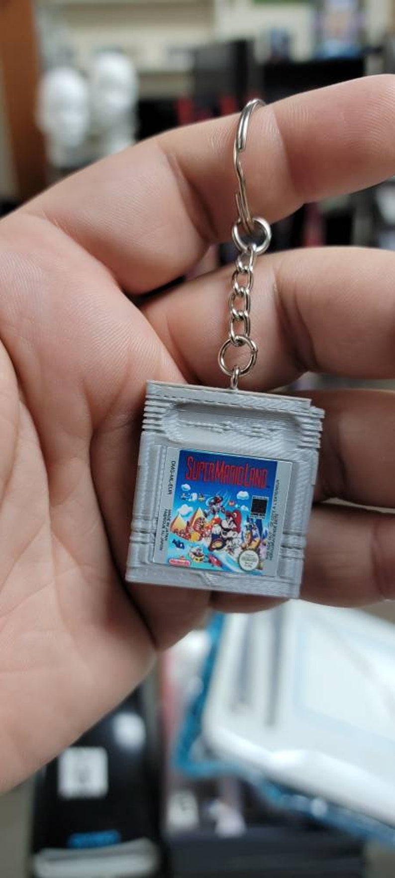 May include: A grey Game Boy Color cartridge keychain with a Super Mario Land game label. The keychain has a silver metal chain attached to a silver metal ring.