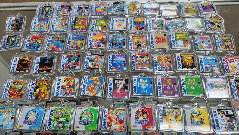 May include: A collection of colourful Game Boy keychains featuring various game titles, including Donkey Kong, Final Fantasy, Zelda, and Pokemon. The keychains are made of clear plastic with a square shape and a metal ring.