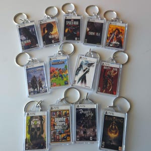 May include: A collection of 12 keychains featuring video game covers. The keychains are made of clear plastic and feature various game titles, including "Spider-Man", "Resident Evil 4", "God of War", "Minecraft", "Final Fantasy VII Remake", "Guilty Gear Strive", "Call of Duty: Modern Warfare", "Grand Theft Auto V", "Demon's Souls", and "Elden Ring".