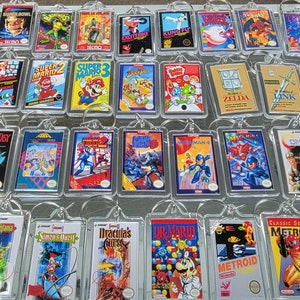 May include: A collection of 25 colorful keychains featuring retro video game cartridges. The keychains are arranged in rows on a glass surface. The cartridges feature popular video game titles such as Super Mario, Zelda, Metroid, and Mega Man.