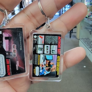 May include: Two clear plastic keychains with images of the Nintendo Entertainment System. One keychain features a pink and gray NES console with the text "ACTION SET" and "Nintendo Entertainment System". The other keychain features a group of people playing a video game with the text "ACTION SET" and "ACTION PACKED ENTERTAINMENT FOR THE ENTIRE FAMILY".