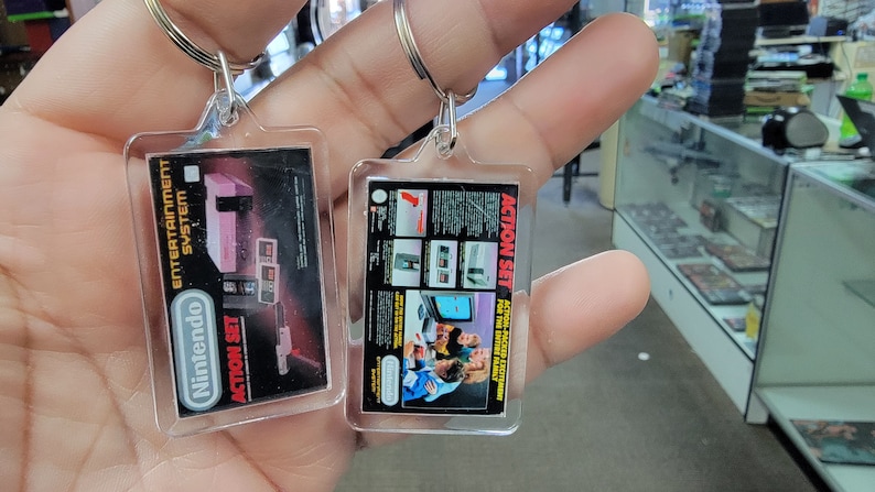 May include: Two clear plastic keychains with images of the Nintendo Entertainment System. One keychain features a pink and gray NES console with the text "ACTION SET" and "Nintendo Entertainment System". The other keychain features a group of people playing a video game with the text "ACTION SET" and "ACTION PACKED ENTERTAINMENT FOR THE ENTIRE FAMILY".