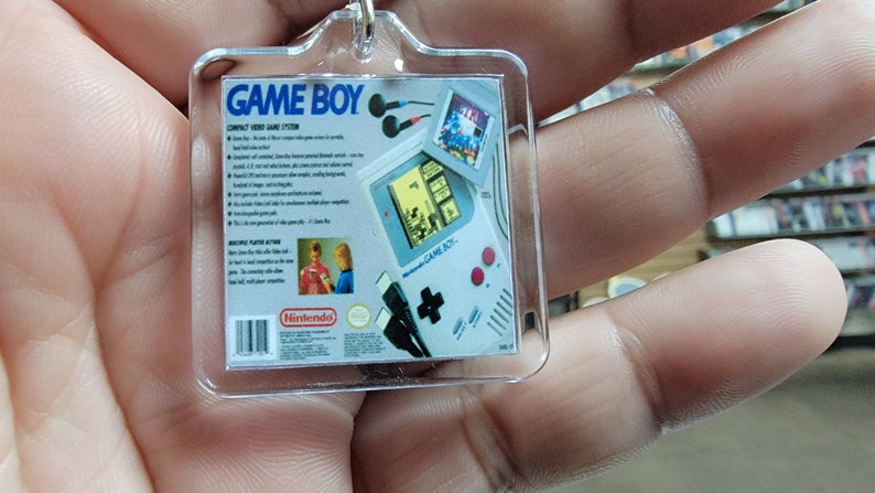 May include: A clear plastic keychain featuring a picture of the back of a Game Boy. The image shows the Game Boy's features, including the screen, buttons, and headphone jack. The text on the back of the Game Boy reads "GAME BOY" and "COMPACT VIDEO GAME SYSTEM".