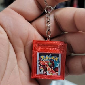 May include: A red 3D printed Game Boy Color keychain with a Pokemon Red game cartridge design. The keychain features the text "Pokemon" and the PEGI rating "3".