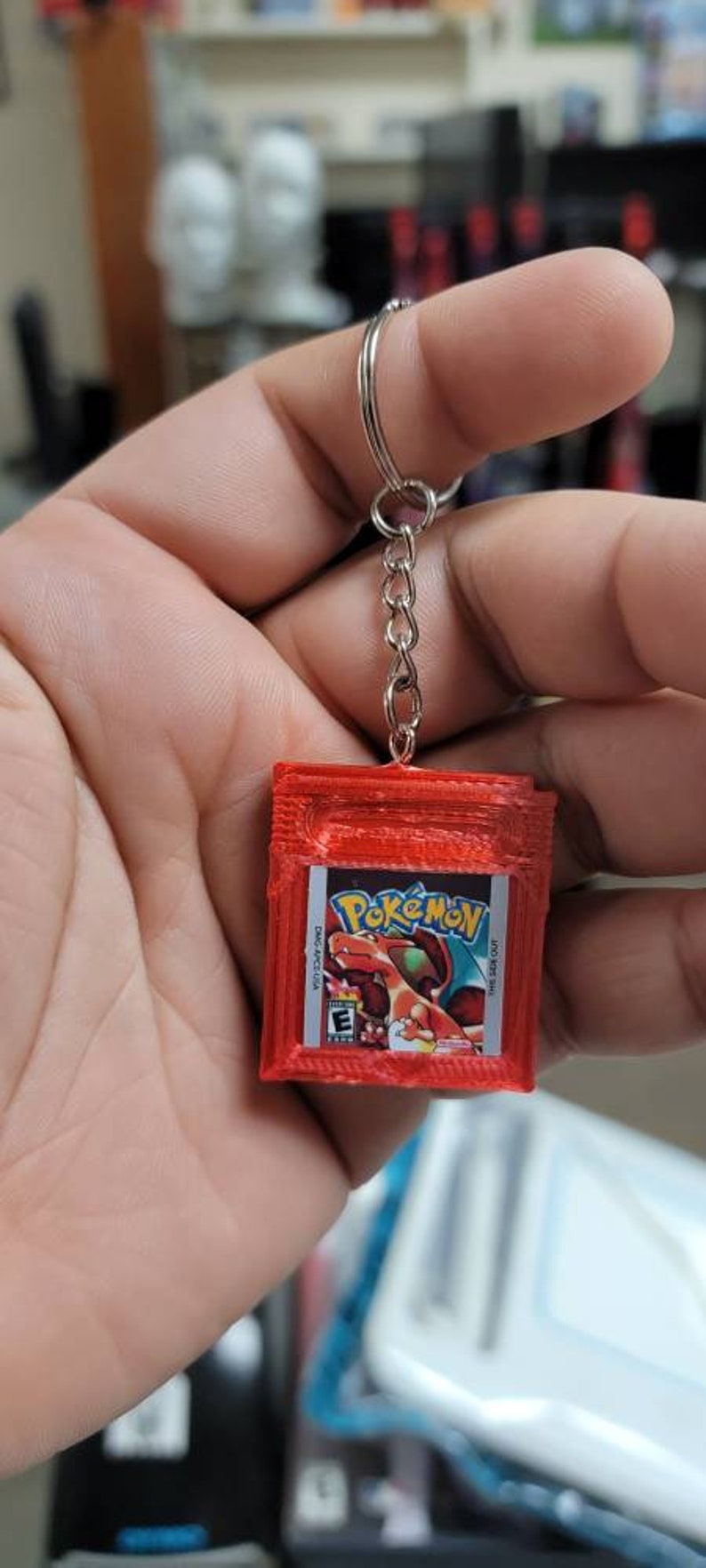 May include: A red 3D printed Game Boy Color keychain with a Pokemon Red game cartridge design. The keychain features the text "Pokemon" and the PEGI rating "3".