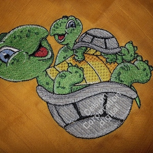 Puede incluir: Diseño bordado de dos tortugas verdes, una llevando a la otra en su espalda. La tortuga más grande tiene una cara sonriente y la tortuga más pequeña tiene la boca cerrada. Las tortugas están sobre un fondo amarillo.