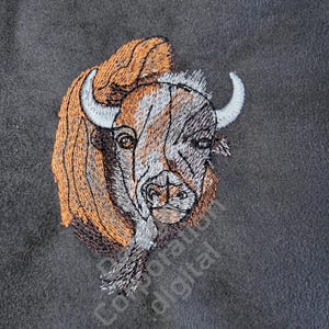 May include: Embroidered image of a brown and white bison head with a long, flowing mane. The bison is facing forward with its head tilted slightly to the side. The background is a dark gray.