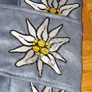 Op de afbeelding: Drie grijze handdoeken van stof met geborduurde witte edelweiss bloemen met gele centra.