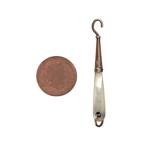 May include: An antique copper-toned button hook with a pearlescent handle, next to a copper coin. The hook has a decorative etched design near the top and a small loop at the bottom. The coin is round with a profile of a person.